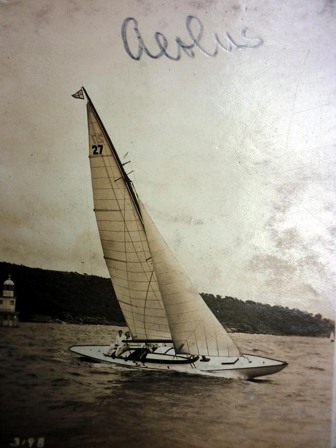 A vintage photo of a Classic Sailing Yacht labelled 27 gliding on the water with two people aboard. Hills and a small building are visible in the background. The word Aeolus is written on the photo’s upper edge.