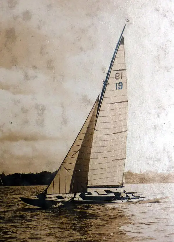 A vintage photograph of a Schärenkreuzer sailboat with the number 19 on its large sail gliding across the water, with land and trees faintly visible in the background under a slightly cloudy sky.