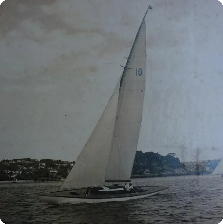 A vintage photograph of a Skerry Cruiser sailboat with the number 19 on its sail, gliding on the water near a shoreline with houses and trees in the background under a cloudy sky.