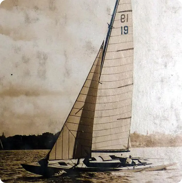A vintage sepia-toned photo of a Schärenkreuzer, its sail marked 19 and 15, gliding gracefully across the water with a faint shoreline and distant trees in the background.