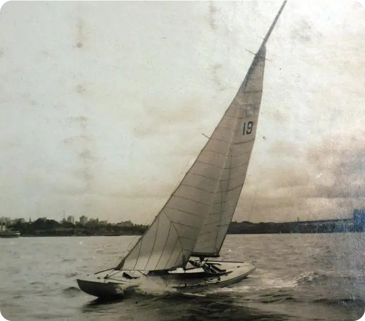 A vintage photo of a Classic Sailing Yacht, sail number 19, heeling as it glides across the water with a person on board; distant shoreline and clouds set the background.