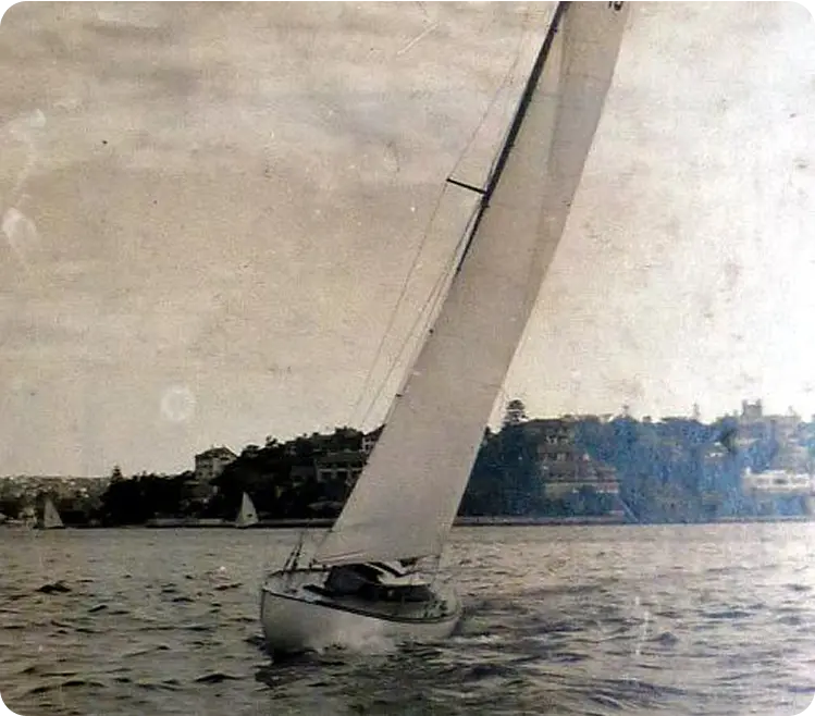 A vintage photograph of a Schärenkreuzer sailboat with a tall mast tilted on the water, sailing near the coast with houses and trees in the background under a partly cloudy sky.
