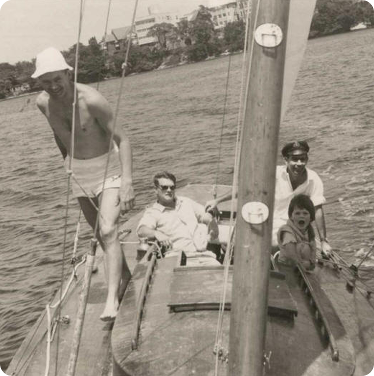 Four people enjoy a day on the water in a classic sailing yacht: a man in swimming trunks stands at the bow, two men sit mid-boat, and a child, mouth open, sits beside them. The Square Metre Yacht glides past trees and buildings on the shore.