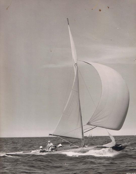 A black and white photo of a Classic Sailing Yacht gliding across the ocean with its large sail billowing in the wind; several people are on board, and the sea and sky extend into the distance.