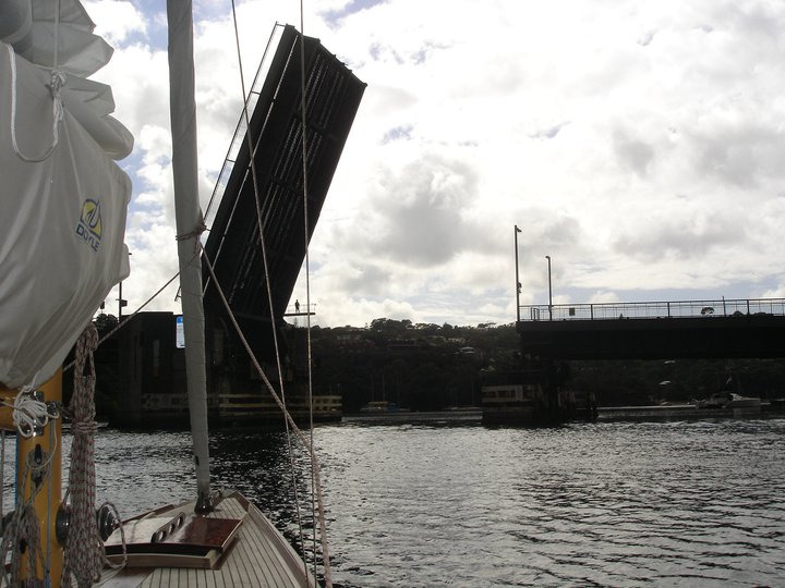 A drawbridge is raised over a body of water, allowing boats such as Schärenkreuzer and other Classic Sailing Yachts to pass underneath. The photo is taken from a sailing boat, with cloudy skies and distant hills in the background.
