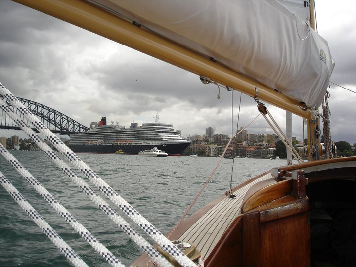 View from a classic sailing yacht of a large cruise ship in the harbour near a city skyline, with the Sydney Harbour Bridge and cloudy skies in the background—an iconic scene for fans of Square Metre Yachts.
