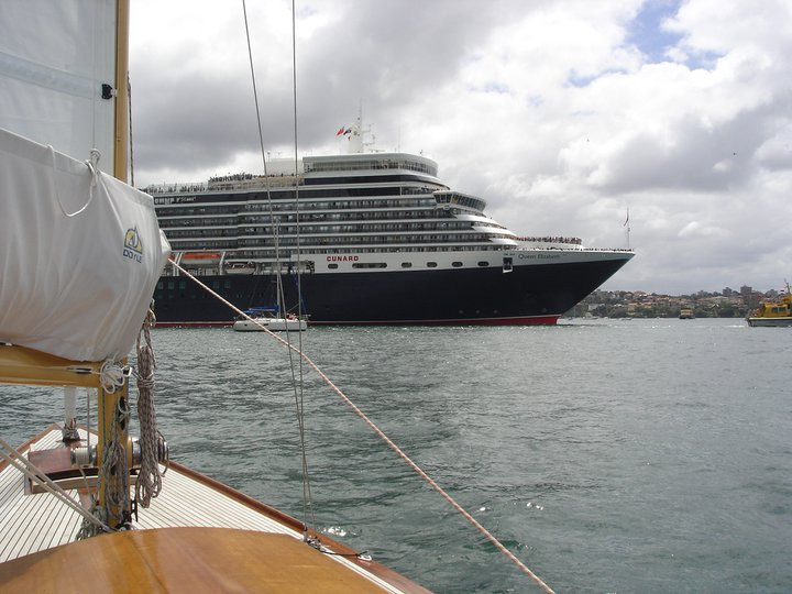 A large black and white cruise ship sails on a cloudy day, viewed from the deck of a classic Square Metre Yacht in the foreground. The water is calm, and a city shoreline is visible in the background.