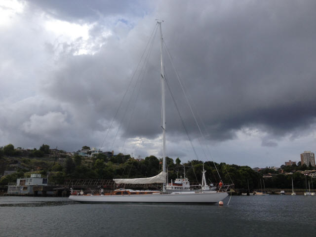A white Skerry Cruiser with its sail down floats on calm water under a cloudy sky, with trees, buildings, and a quay in the background.