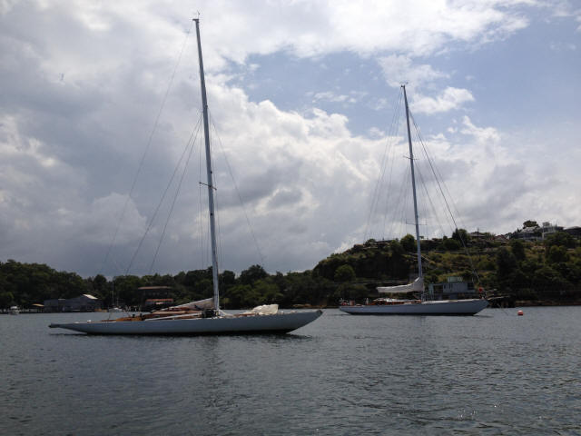 Two Classic Sailing Yachts anchored on calm water near a green, tree-covered shoreline, with houses on a hill and a partly cloudy sky overhead.