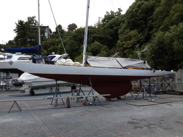 A Square Metre Yacht with a red keel is propped up on stands in a boatyard, surrounded by trees and other boats, with supplies and a tarpaulin covering part of the deck.