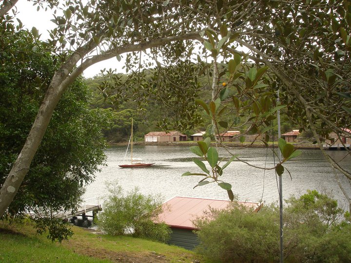 A small Classic Sailing Yacht glides on a calm river, framed by tree branches. Boathouses and forested hills line the opposite bank, with a jetty and green-roofed building in the foreground.