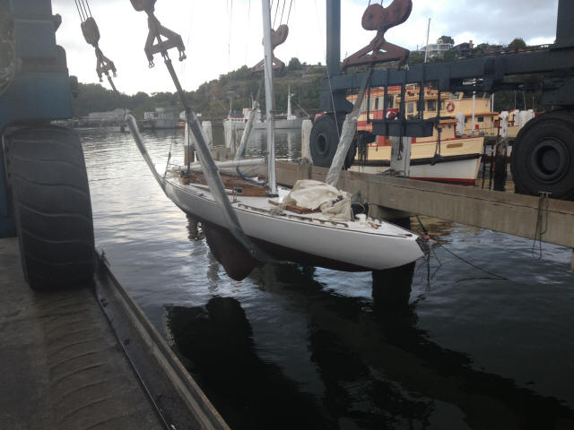 A white sailboat, reminiscent of Classic Sailing Yachts, is being lifted out of the water by a large boat hoist at a marina, with another boat and trees visible in the background.