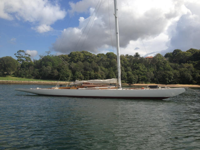 A sleek white Square Metre Yacht with its sails down is anchored on calm water near a forested shoreline under a partly cloudy sky.