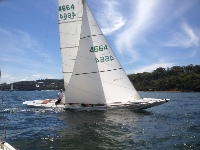 A white Square Metre Yacht with the number 4664 sails on a blue, slightly wavy body of water near a forested shoreline under a partly cloudy sky. A person sits at the stern of this classic sailing yacht, steering.