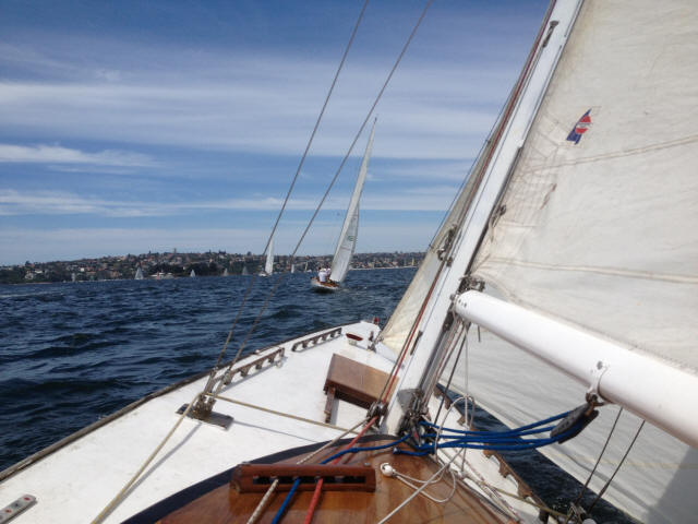 View from the deck of a Classic Sailing Yacht on the water, sails up and ropes visible; another sailboat is seen ahead under a blue sky with light clouds, with a distant shoreline in the background.
