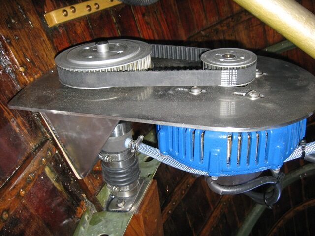 A blue electric motor is mounted on a wooden surface, connected to a metal plate with two pulleys and a black belt—similar to precision equipment found on Classic Sailing Yachts such as Square Metre Yachts or Schärenkreuzer.