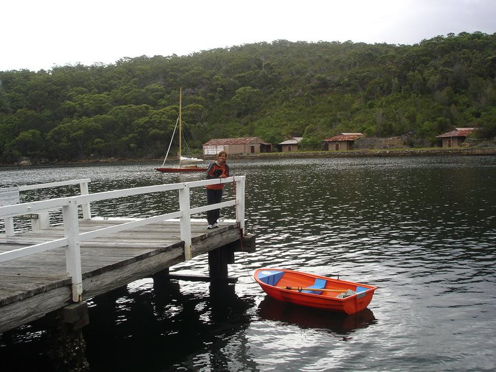 A person stands on a wooden jetty above a calm body of water, with a small orange rowing boat tied to the jetty. A classic sailing yacht and several cabins are visible on the green, tree-covered shore in the background.