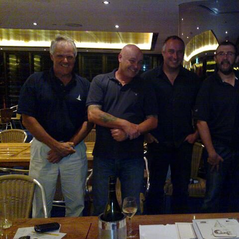 Four men are standing and smiling indoors at a restaurant, wearing dark shirts. On the table in front of them, beside a wine bottle and papers, you’ll find talk of Classic Sailing Yachts in the relaxed social setting.
