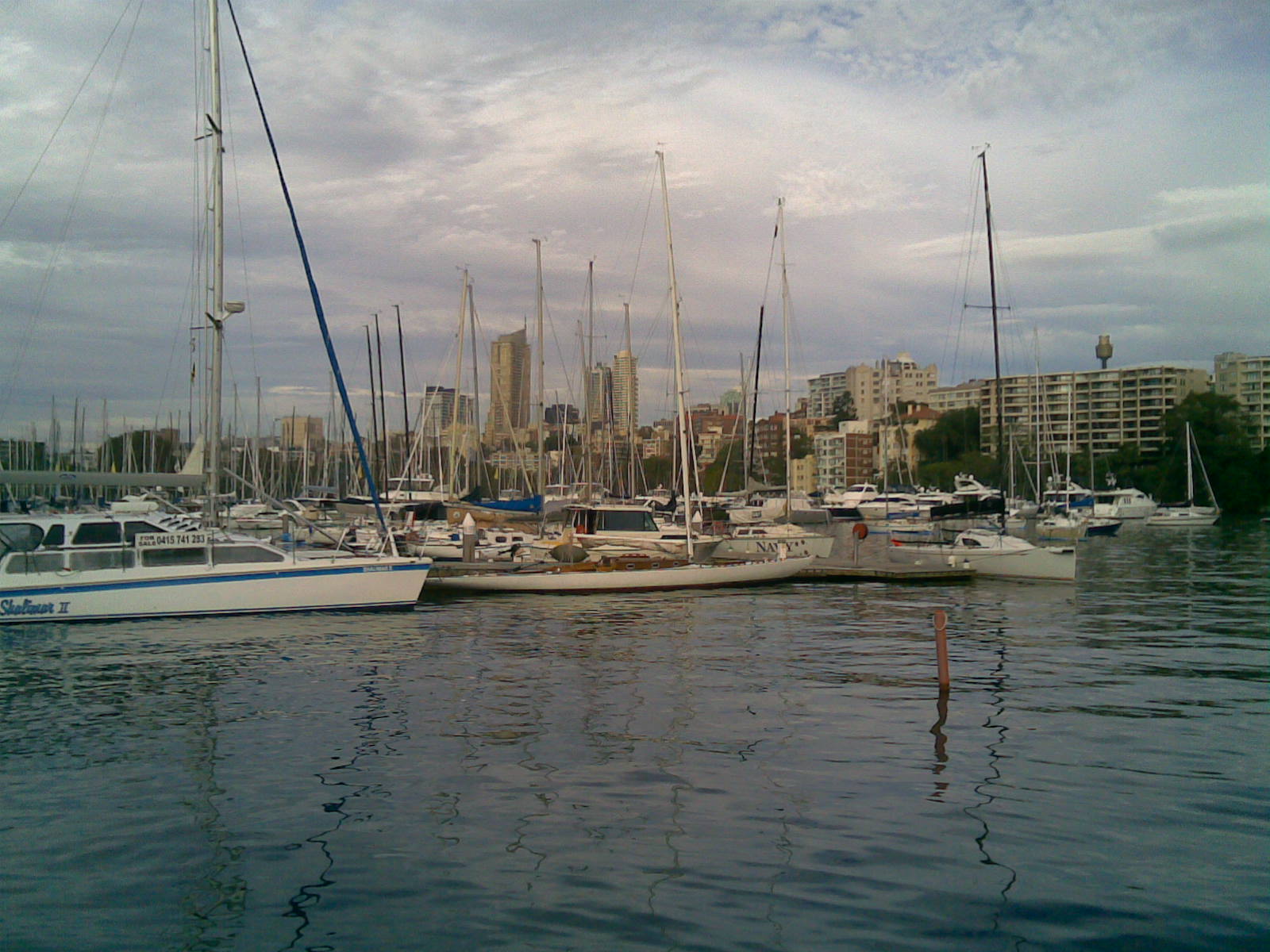 Several Square Metre Yachts and classic sailing yachts are moored in a marina, with tall buildings and flats in the background under a cloudy sky. The serene scene is beautifully reflected on the calm water below.