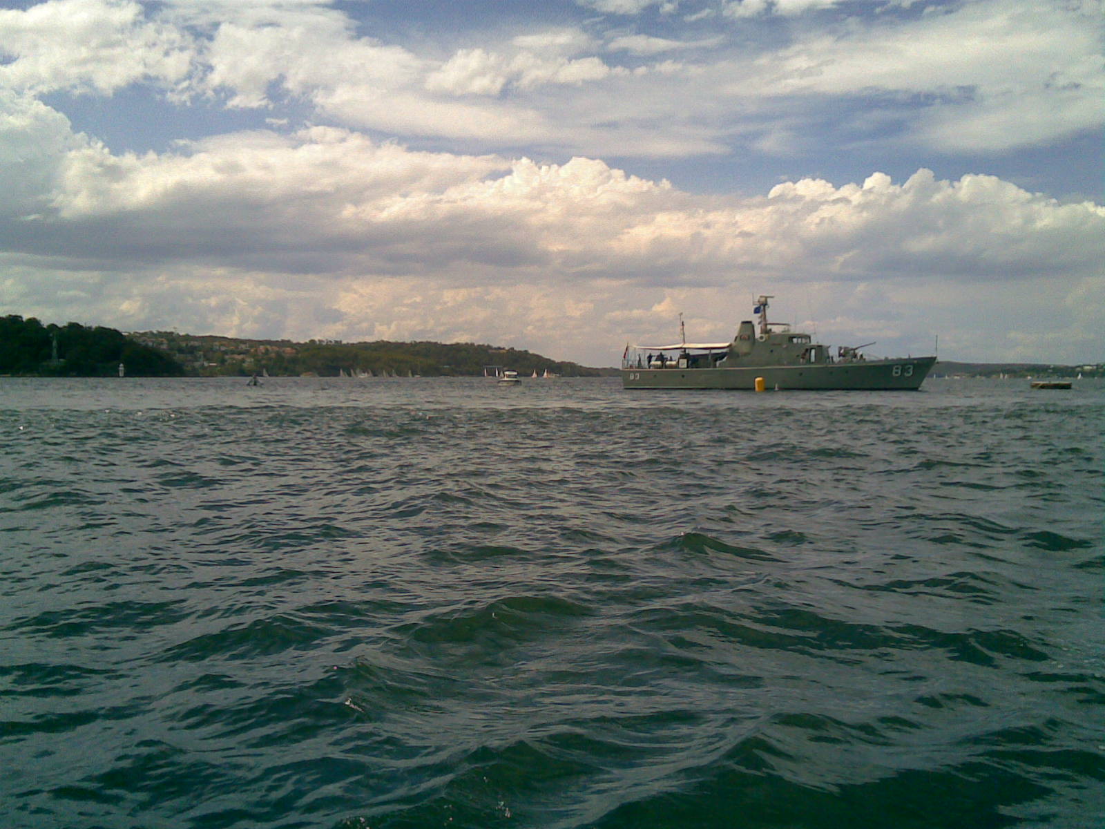 A military ship sails on a large body of water under a partly cloudy sky, with classic sailing yachts and small boats visible near the distant shoreline in the background.