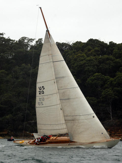 A classic Square Metre Yacht with white sails marked US 20 glides on choppy water near a forested shoreline under an overcast sky. Several people are visible on board, adjusting the sails.