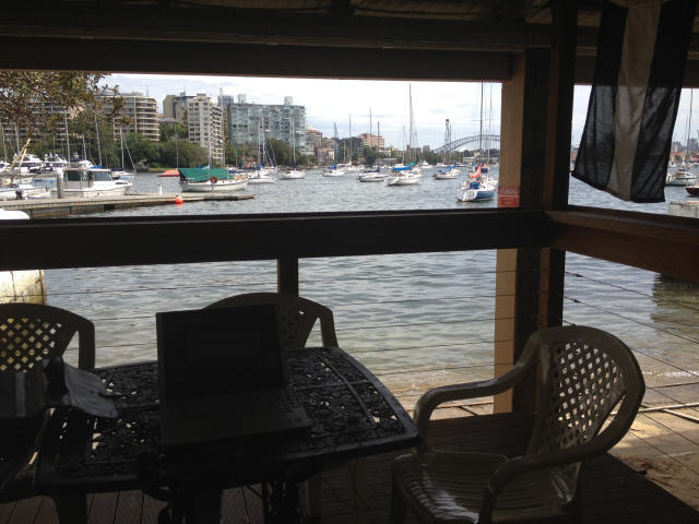A shaded patio with a table, laptop, and chairs overlooks a marina featuring elegant Skerry Cruisers moored on the water. City buildings and a bridge rise in the background beneath a partly cloudy sky.