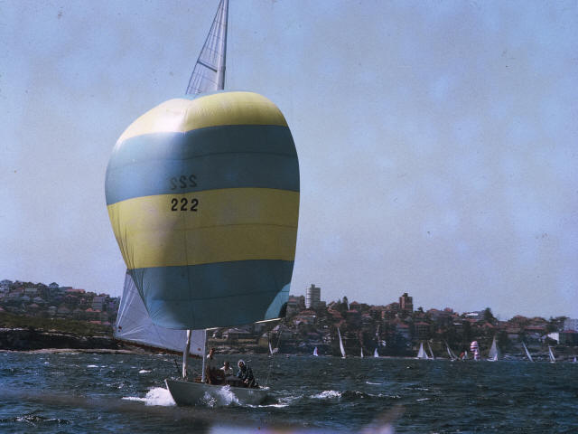 A classic sailing yacht with a large yellow and blue spinnaker marked SSS 222 sails on open water, with a coastline and more boats visible in the background under a clear sky.