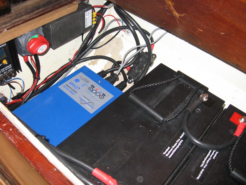 A close-up view of batteries and electrical components, including cables and a blue battery charger, arranged inside a compartment with a wooden cover—typical of the meticulous setups found on Classic Sailing Yachts.