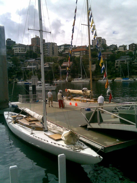 Several people stand on a quay next to classic sailing yachts, one of which is decorated with colourful flags. The scene overlooks calm water with more boats and hillside buildings in the background.