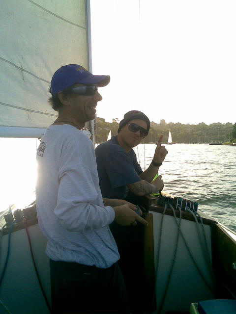 Two people on a sailboat, both smiling and wearing sunglasses. One is also wearing a beanie and making a peace sign. Classic Sailing Yachts drift nearby, with trees and other boats in the background.