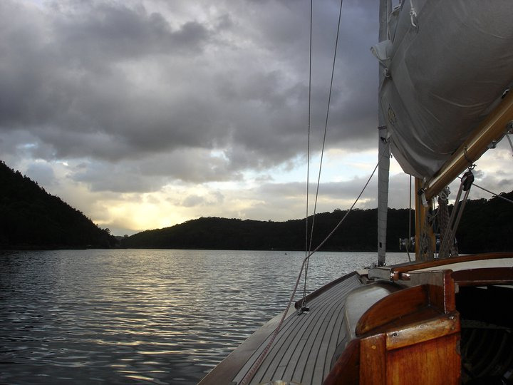A view from a classic Schärenkreuzer sailboat on calm water, with forested hills in the distance under a cloudy sky and a hint of sunset light peeking through.