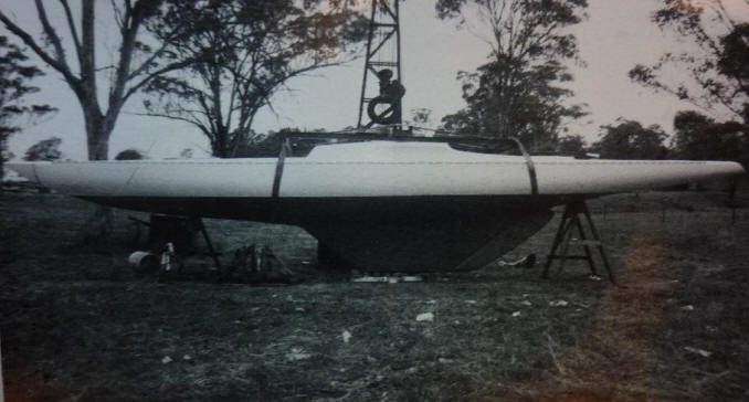 A black and white photo of a classic sailing yacht on land supported by stands, with a person sitting on top near a metal frame, surrounded by trees and grassy ground.