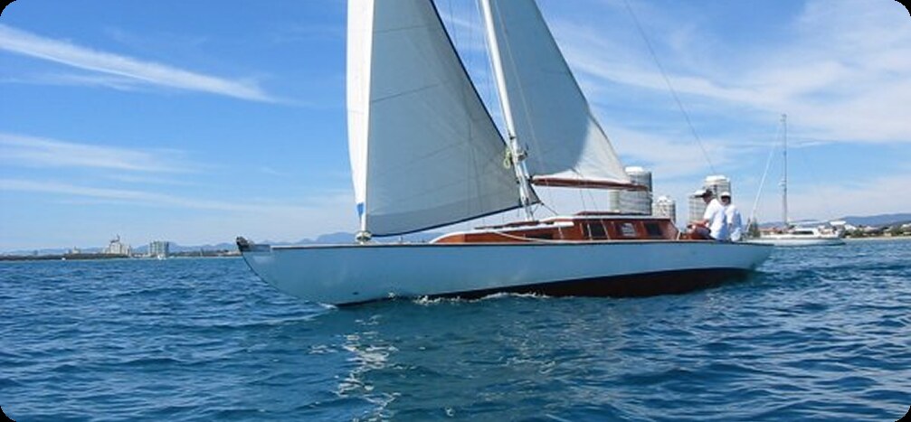 A person sails a small white Schärenkreuzer with blue trim on a calm body of water under a clear blue sky, with buildings and other boats visible in the background.