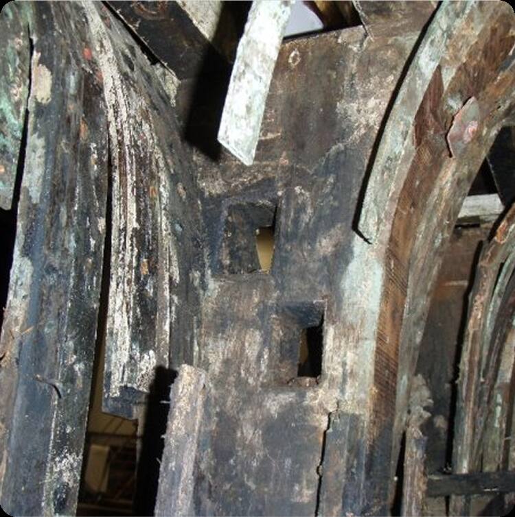 A close-up of weathered, decaying wooden beams with square cut-outs, reminiscent of old Skerry Cruisers; some beams show greenish patches and splitting from moisture exposure, evoking the history of Square Metre Yachts.