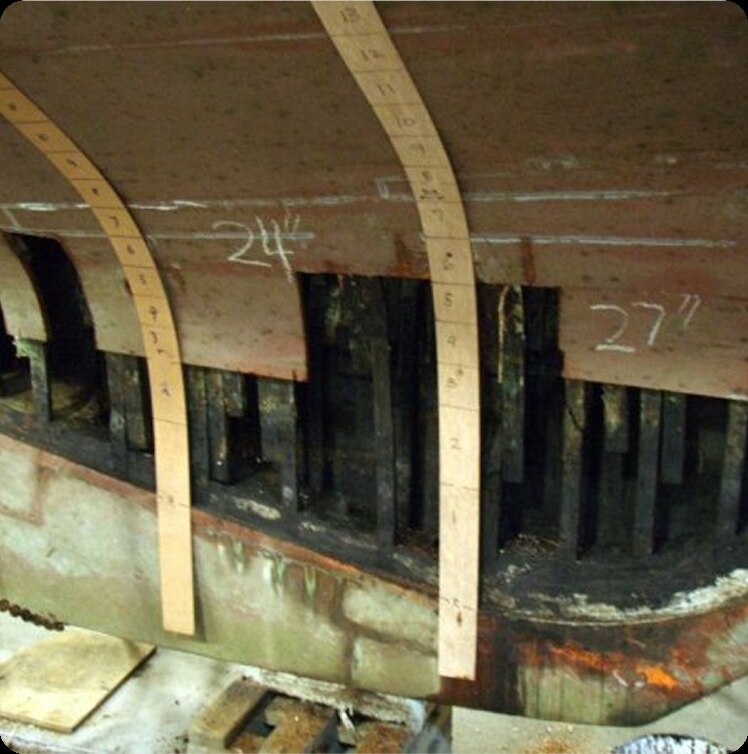 Close-up of a Square Metre Yacht hull under repair, showing exposed wooden frames and ribs. Two measuring strips and chalk markings reading 24½ and 27” are visible on the hull surface.