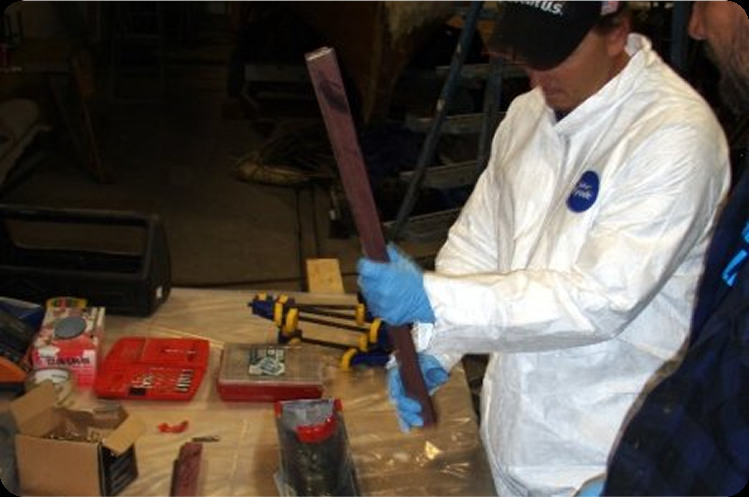 Two people work at a cluttered table covered with tools and supplies. One, wearing a white protective suit and blue gloves, holds a long, rectangular object whilst the other stands nearby, possibly preparing parts for Classic Sailing Yachts or Schärenkreuzer repairs.