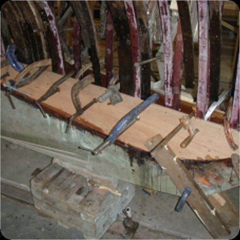 Wooden boat under repair with multiple clamps securing new planks to the frame; ribs of this classic sailing yacht are visible in the background.