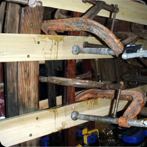 Several large cramps hold wooden boards together, with some water dripping from the boards—perhaps materials being prepared for restoring Schärenkreuzer or classic sailing yachts in a woodworking project.