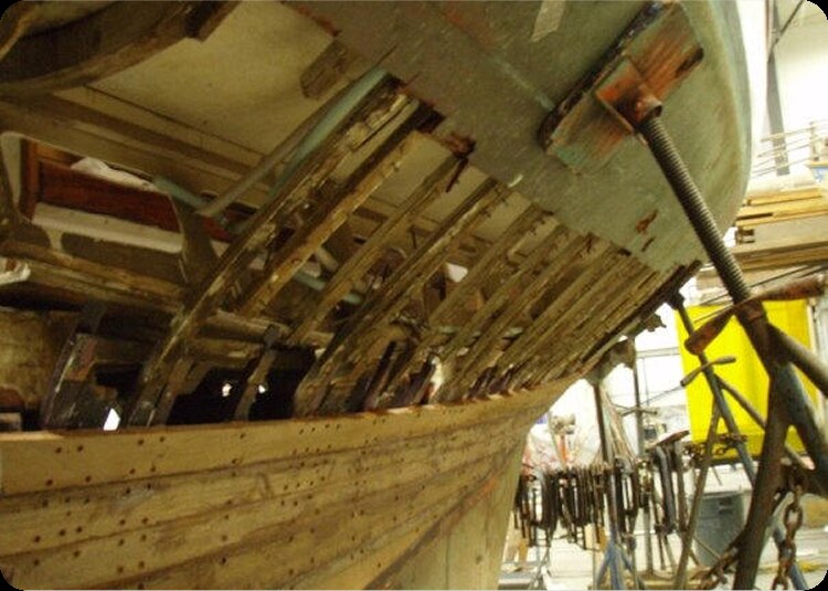 A close-up view of a wooden Classic Sailing Yacht under restoration, showing exposed ribs and clamps holding pieces of wood in place in a workshop.