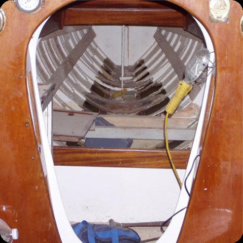 View inside a wooden boat hull—possibly a Skerry Cruiser or other classic sailing yacht—showing exposed internal ribs and frame; a portable work light with a yellow lead hangs on the right, tools or equipment visible at the bottom of the frame.
