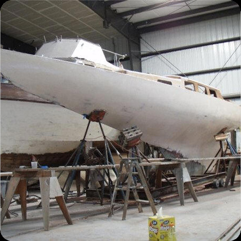 A large white Classic Sailing Yacht is being repaired or constructed indoors, supported by stands and wooden planks. Various tools and equipment are scattered around, including a yellow box in the foreground.