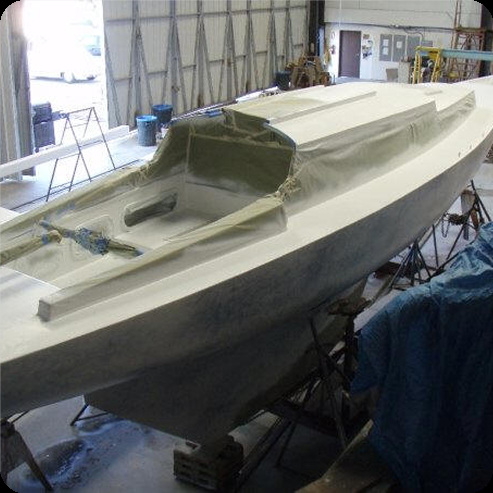 A white Schärenkreuzer sailboat hull is under construction inside a workshop, supported by stands. The deck is partially covered with protective material, and tools and equipment are visible in the background.