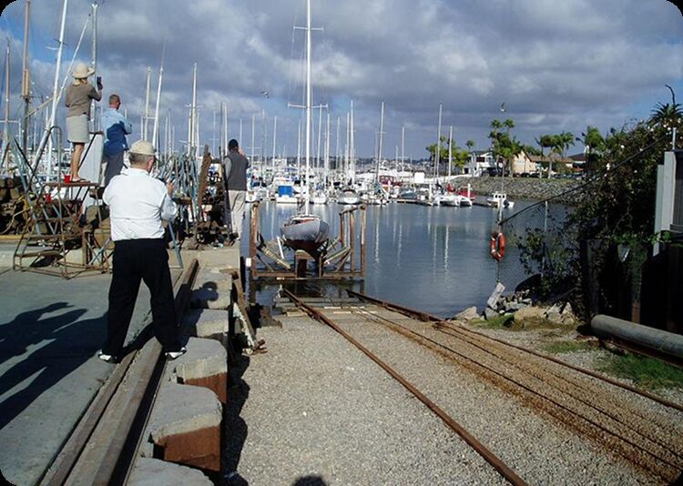 People stand near slipways and railway lines beside a marina, taking photos of a classic Schärenkreuzer sailboat being launched into the water. Numerous Square Metre Yachts are moored in the background under a partly cloudy sky.