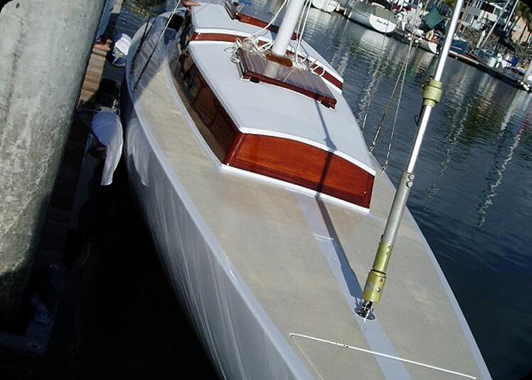 A sleek white Square Metre Yacht with a polished wooden cabin is moored at a marina. A person stands on the quay beside the boat, with other boats and waterfront buildings visible in the background.