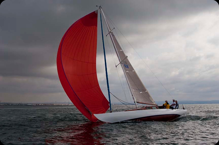 A graceful Square Metre Yacht with a large red spinnaker sail glides across the water under a cloudy sky. Several people are visible on board, leaning to one side as the boat tilts in the wind.