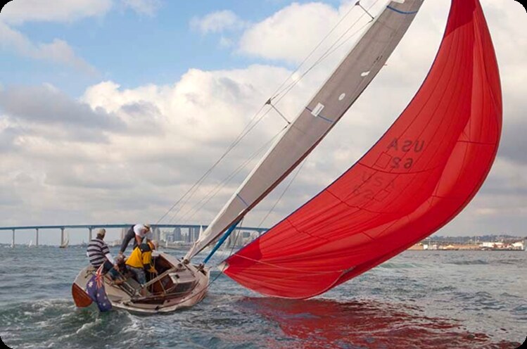 Three people sail a small Schärenkreuzer with a large, billowing red sail on the water, leaning to one side. A city skyline and a long bridge are visible in the background under a partly cloudy sky.