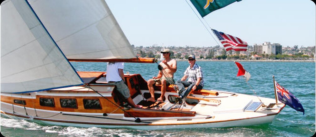 Three people sail a classic wooden Square Metre Yacht on blue water near a city shoreline. The boat displays several flags, including the American flag, as they enjoy the sunny, clear weather.