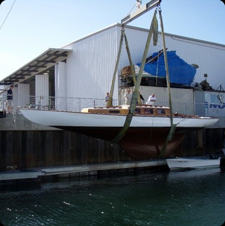 A Schärenkreuzer sailboat is being lifted by a crane with green straps beside a quay near a large white building. Several people are standing on the quay observing the process.