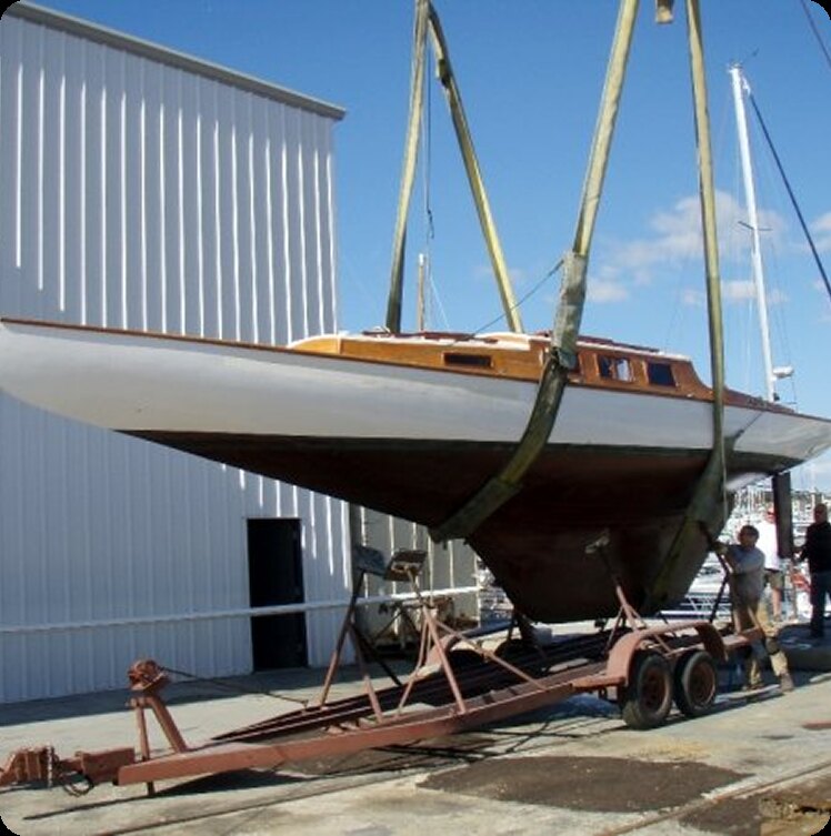 A Square Metre Yacht is being lifted by straps attached to a crane, hovering above a trailer on a quay. Two people stand nearby, and a white building and other boats are visible in the background under a blue sky.