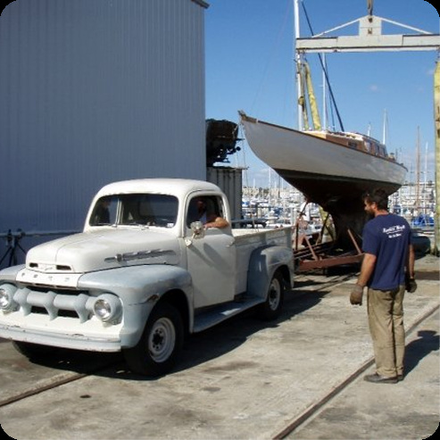 A vintage white pickup truck is parked near a marina whilst a large wooden Schärenkreuzer sailboat is being hoisted by a crane. A man in a blue shirt and gloves stands watching. Sailboats, including other Skerry Cruisers, are visible in the background.